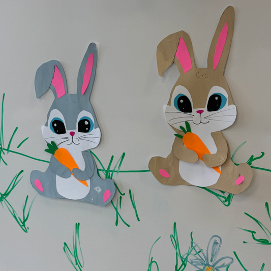A gray bunny and a light brown bunny both holding carrots on white background with hand=drawn grass around them